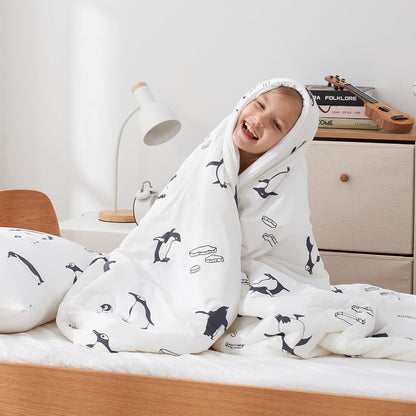 Child wrapped in a white cooling comforter with antarctic penguin prints, sitting on a bed.#color_Antarctic Penguin
